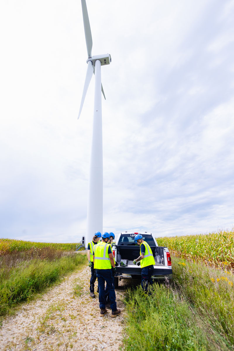 Technicians inspect equipment beside a wind turbine and pickup truck.